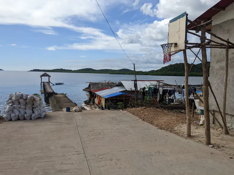Concrete pier extending into the sea beside houses with tin roofs and a makeshift basketball hoop.