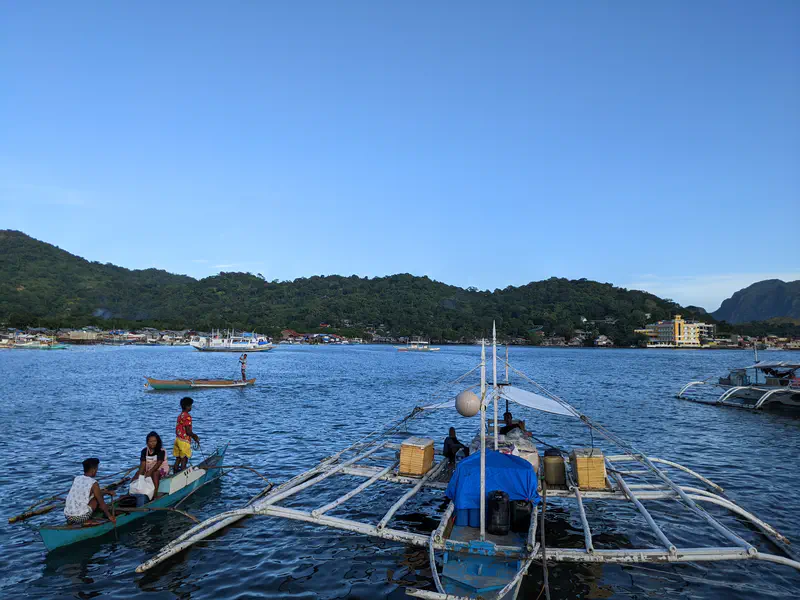Small fishing boats with people aboard on the water, with a larger outrigger boat in the foreground and green hills in the background.
