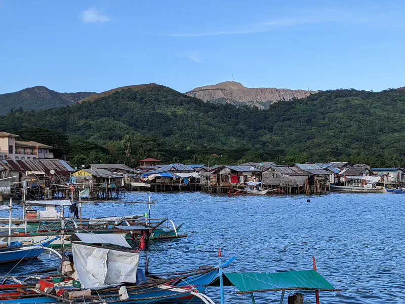 Fishing boats docked near stilt houses over water with green hills and mountains in the background.
