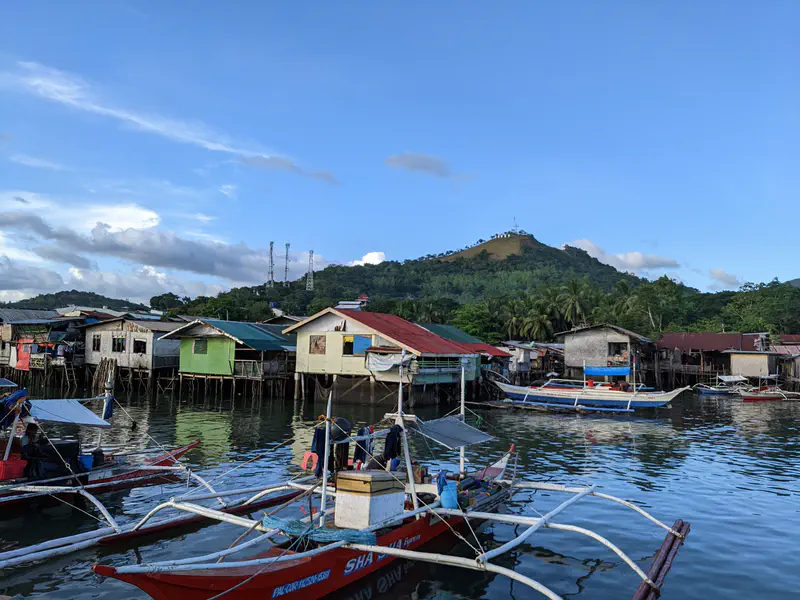 Colorful stilt houses along the water with traditional fishing boats docked, and a hill with antennas in the background.