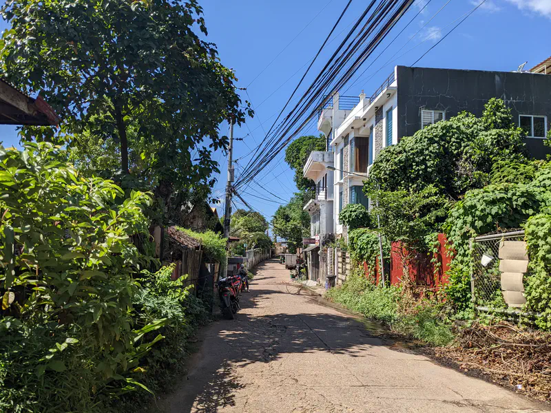 Sunny narrow street lined with trees, motorbikes, and residential houses with many overhead power lines.