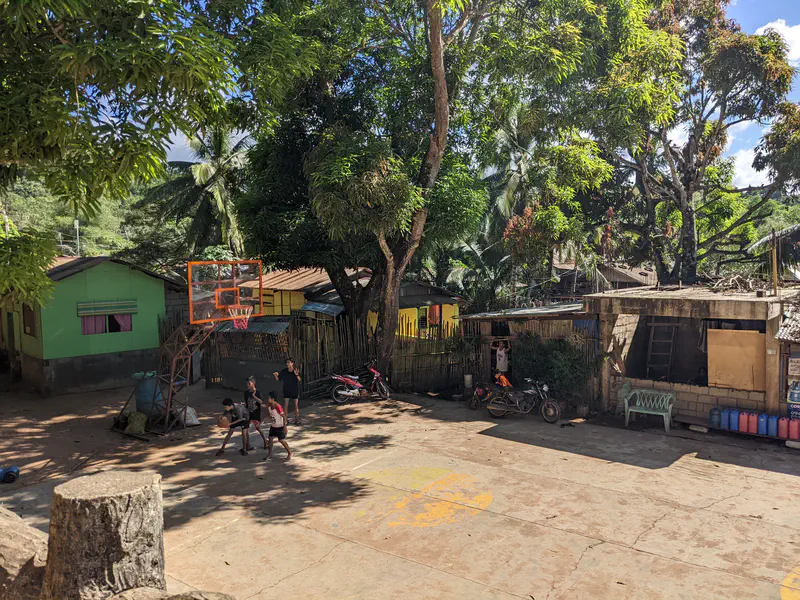 Children playing basketball on an outdoor court beside small houses and trees.