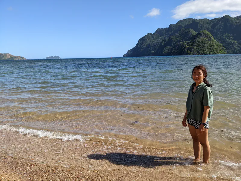 Woman standing at the edge of the sea with clear water and sandy shore, smiling with green hills and rocky cliffs in the background.
