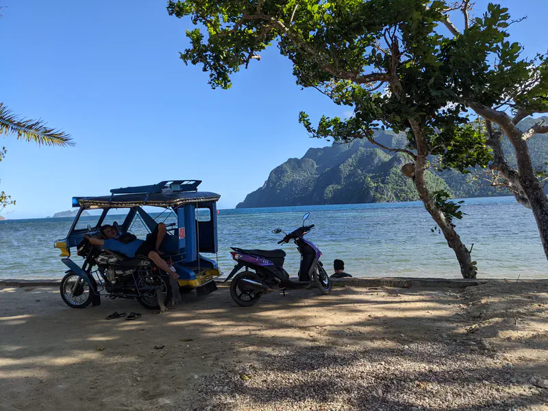 Man resting on a tricycle beside a motorbike under trees by the sea with mountains in the distance.