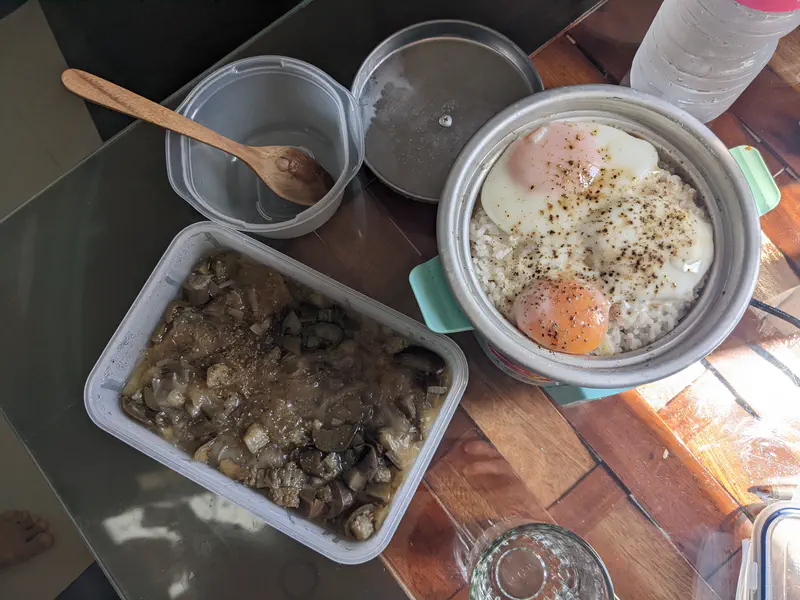 Meal of rice with fried eggs and a container of cooked vegetables on a table.