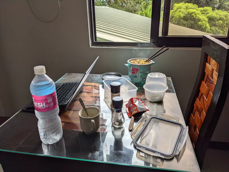 Table with a laptop, bottled water, and a steaming bowl of noodles beside condiments.