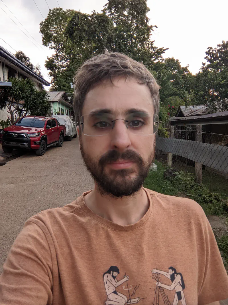 Man with glasses and beard taking a selfie on a village street with houses and a red car.