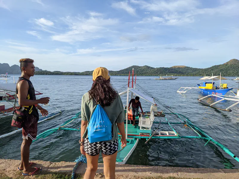 Two people standing near a bangka boat at the shore with other boats and green hills in the background.