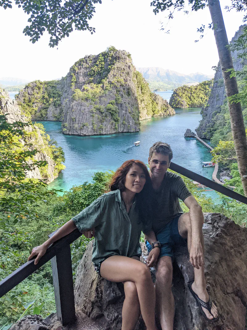 Two people sitting on a rock with a view of turquoise water and limestone cliffs in the background.