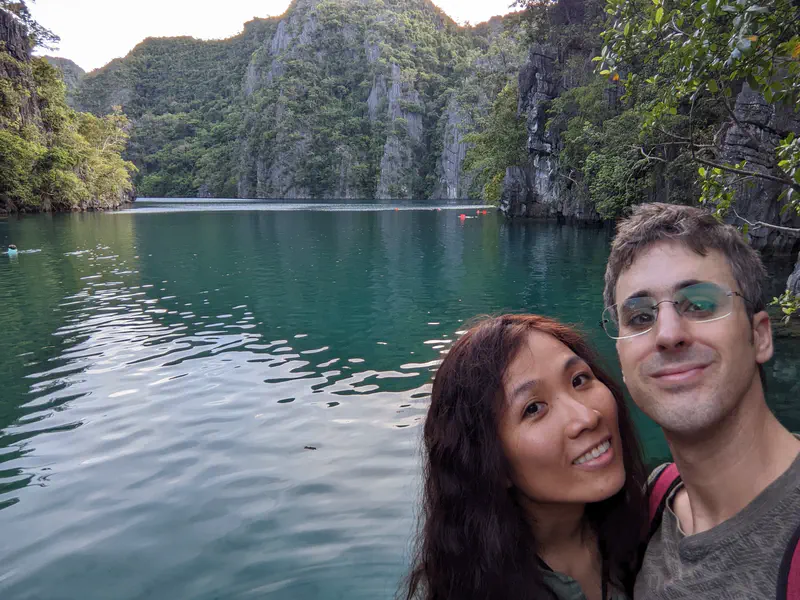 Two people taking a selfie by a calm turquoise lagoon surrounded by limestone cliffs and greenery.