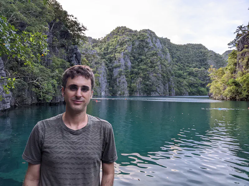 Man standing in front of a turquoise lagoon with limestone cliffs and lush trees in the background.