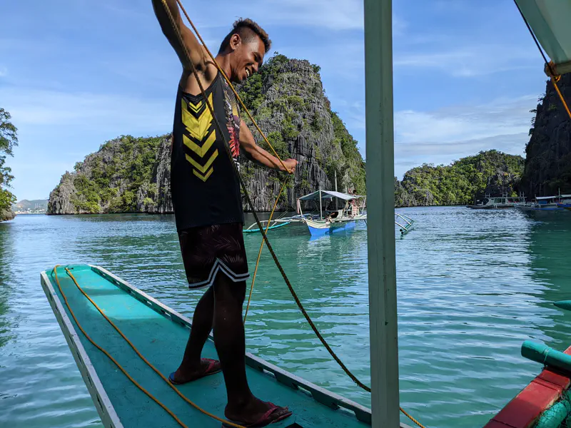 Man holding a rope on a bangka boat in turquoise water surrounded by limestone cliffs.
