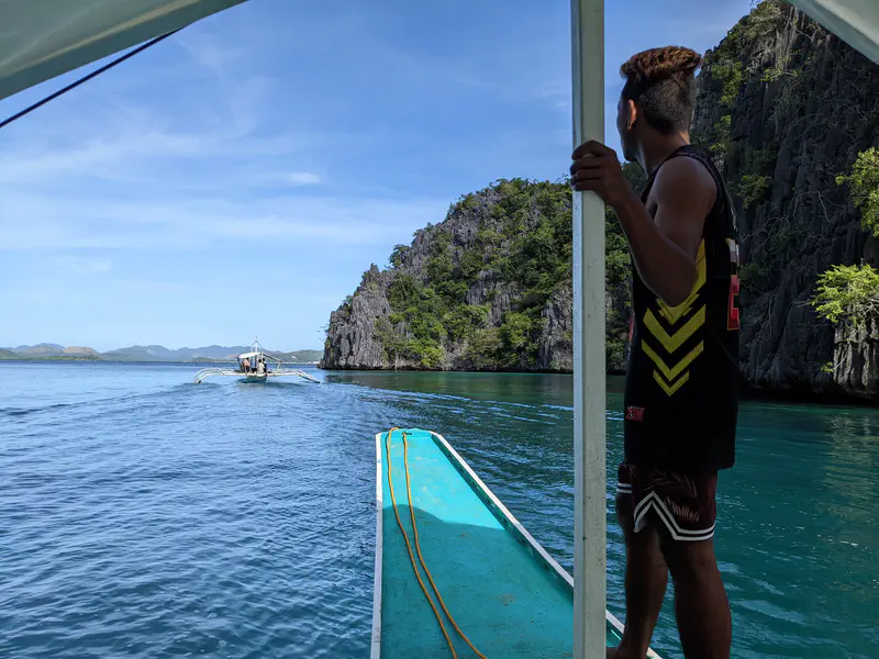Man standing on a bangka boat looking out at another boat on turquoise water between limestone cliffs.