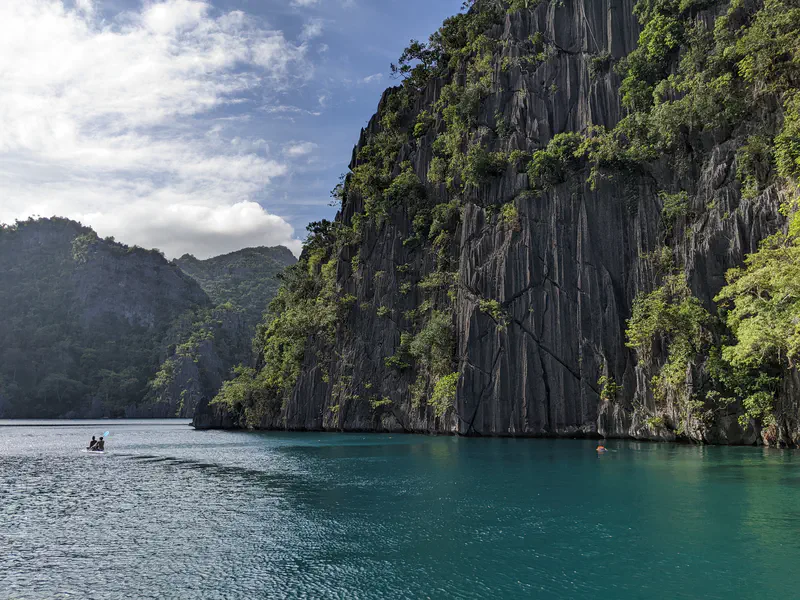 Cliffs rising above a turquoise lagoon with people swimming and kayaking in the distance.