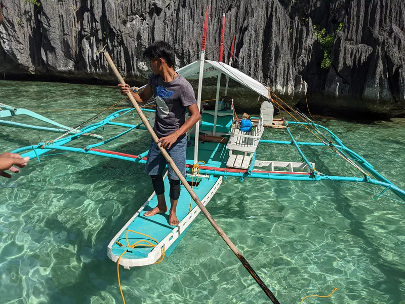 Man standing on the edge of a bangka boat with a wooden pole in clear turquoise water near limestone cliffs.