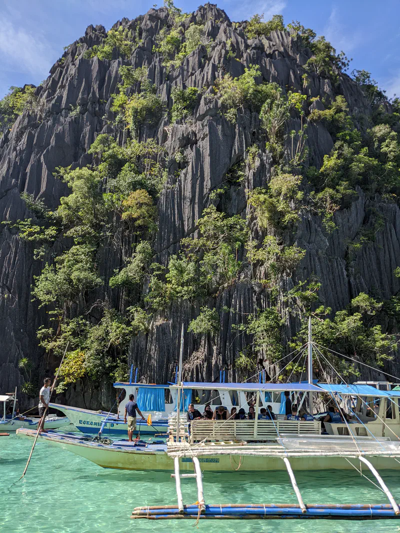Tour boats with passengers floating on turquoise water at the base of a tall limestone cliff with greenery.