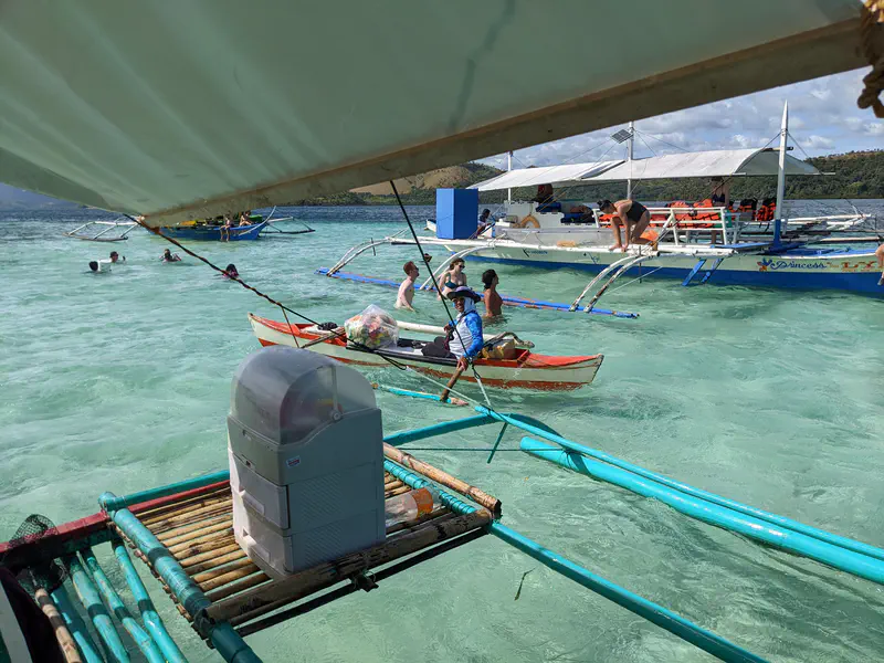 People swimming and small boats floating in shallow turquoise water near larger tour boats.