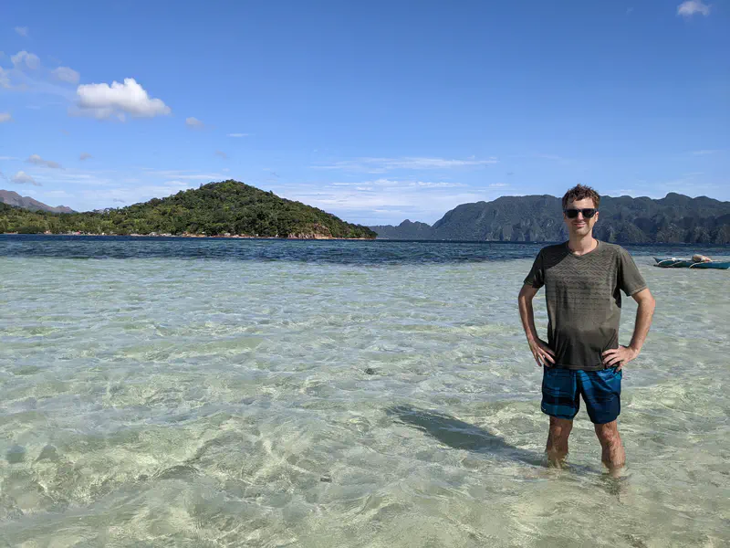 Man standing in shallow clear water with green islands and mountains in the background.