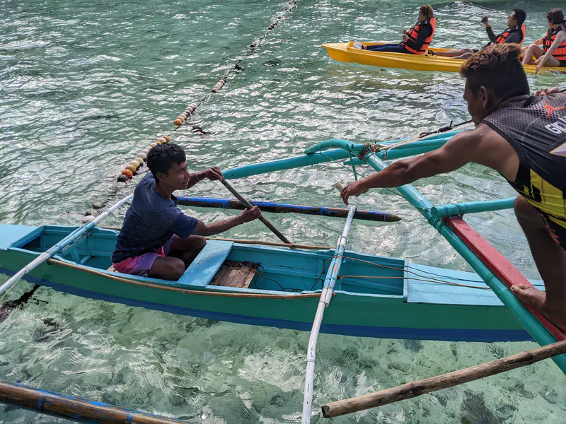Man in a small wooden boat being assisted by another man while people in kayaks float nearby.