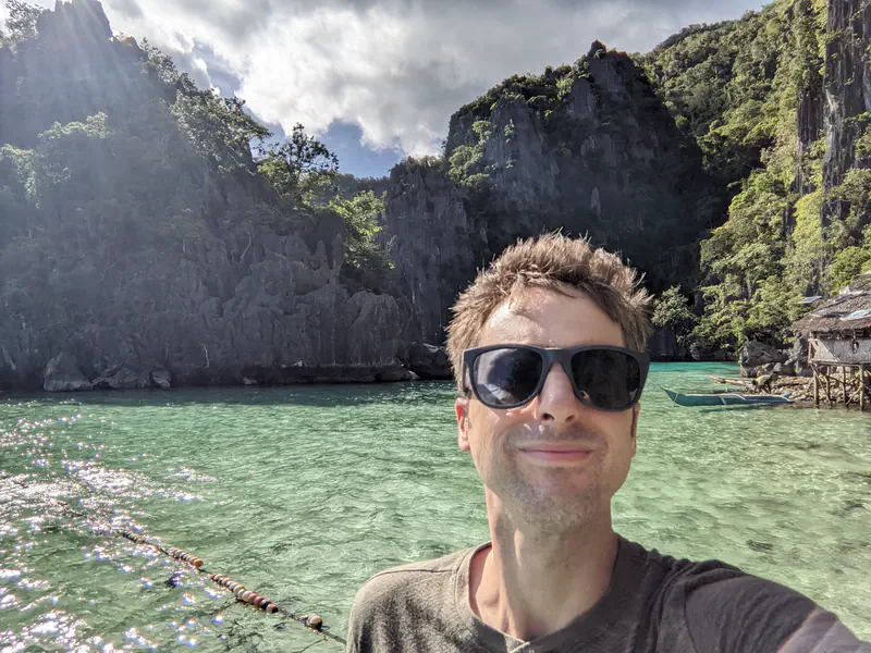 Man taking a selfie by clear turquoise water with limestone cliffs and trees in the background.