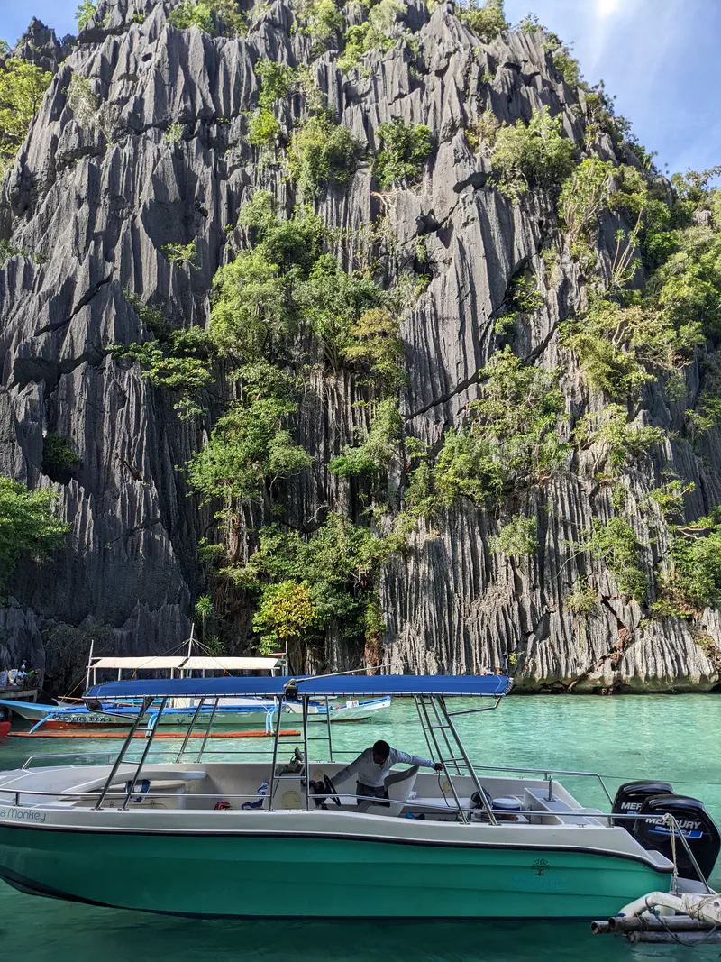 Motorboat floating on turquoise water in front of a tall limestone cliff with greenery.