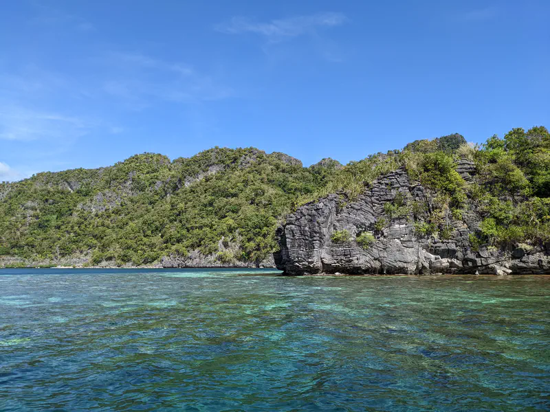 Rocky island with lush green vegetation surrounded by clear turquoise water under a blue sky.