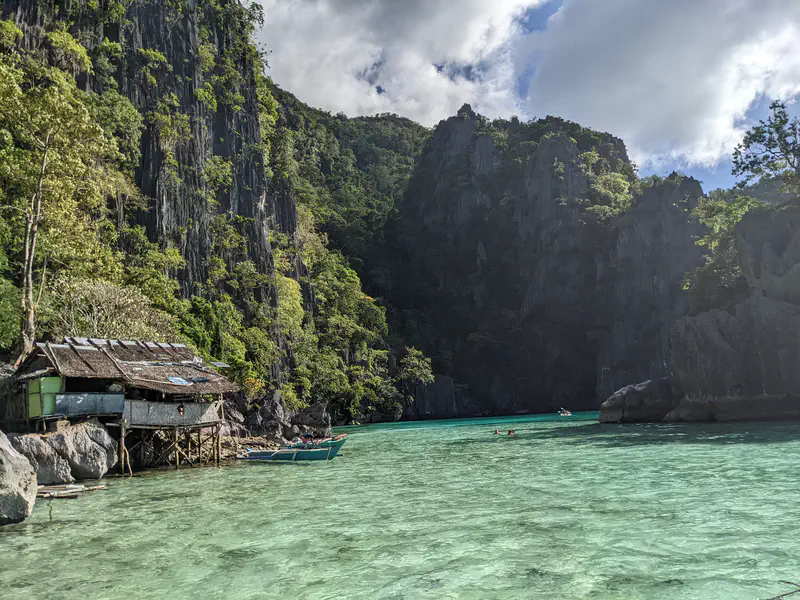 Thatched hut on stilts by turquoise water with limestone cliffs and swimmers in the distance.