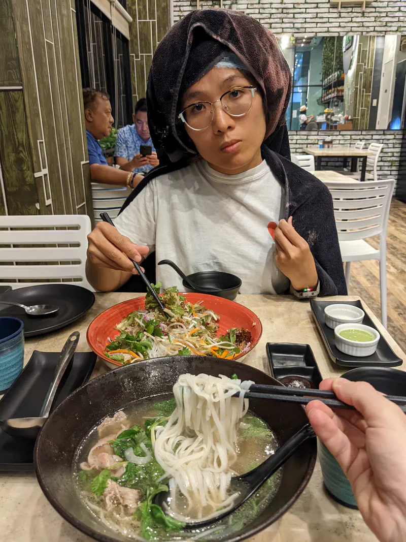 Woman with a towel on her head eating noodles at a restaurant table with pho and salad dishes.