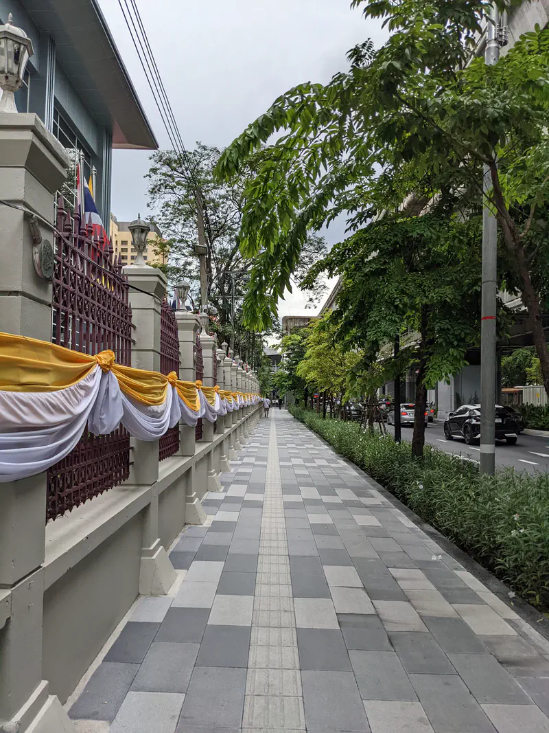 Tree-lined sidewalk in Bangkok with decorative yellow and white fabric draped on a fence, extending into the distance.