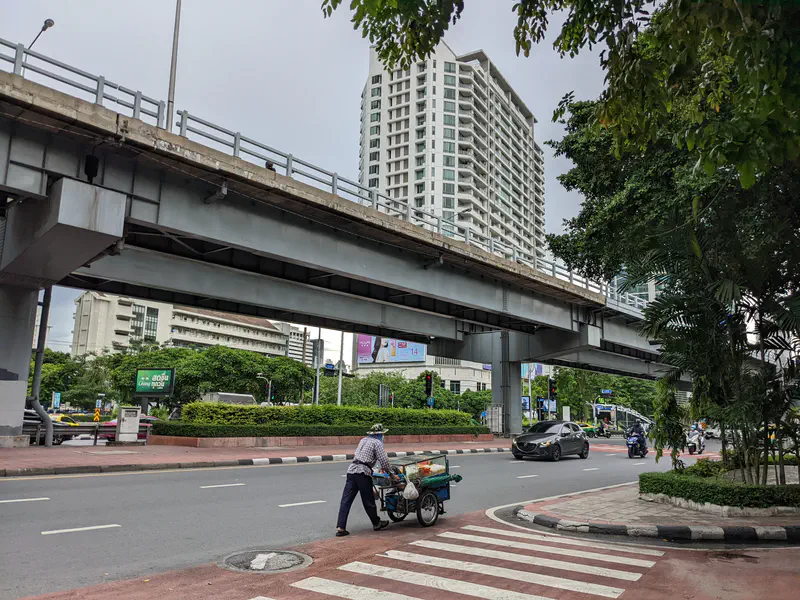 Man pushing a cart across a crosswalk under an elevated roadway in Bangkok, with tall buildings and traffic nearby.