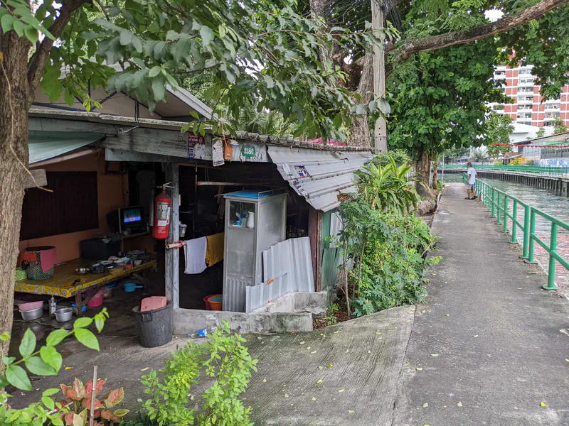 Small riverside dwelling in Bangkok with an open kitchen area, fire extinguisher, and pathway along the canal.