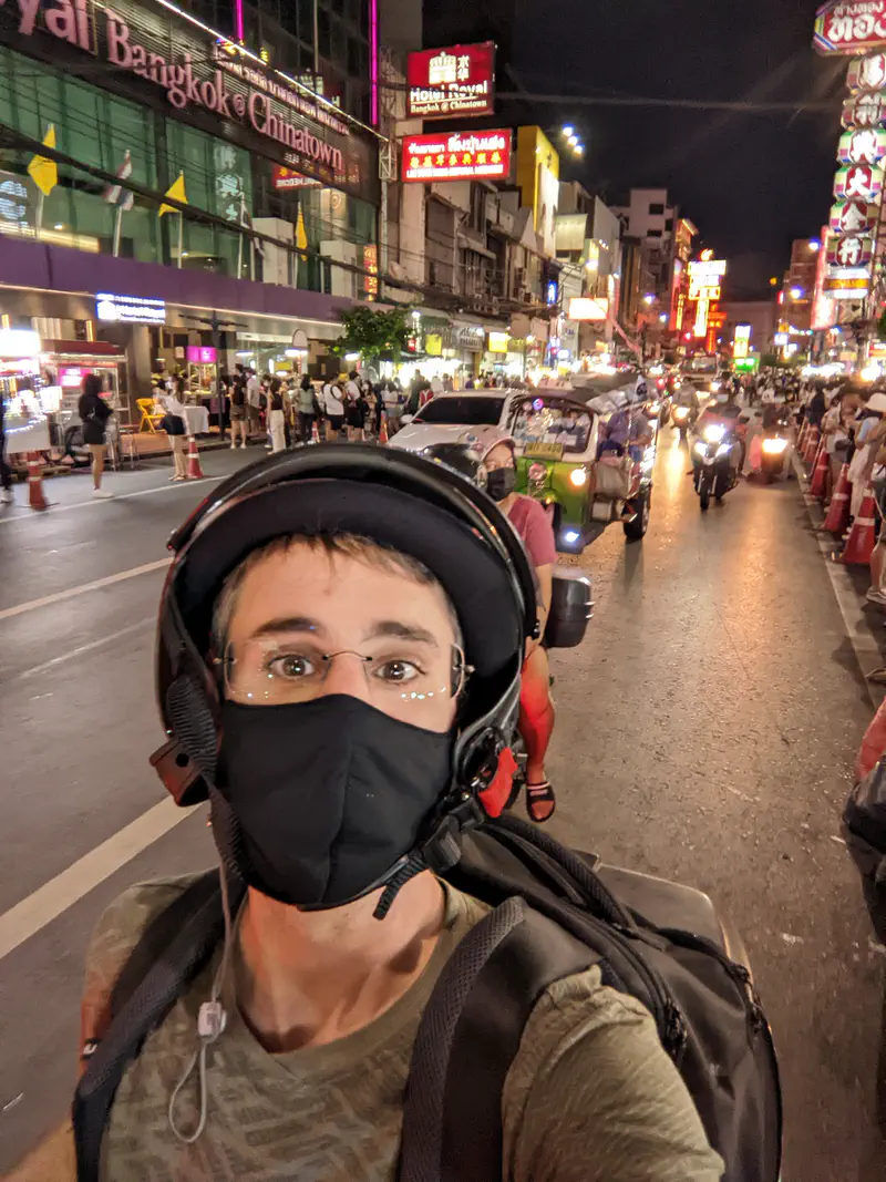 Person wearing a helmet and mask taking a selfie on a busy night street in Bangkok’s Chinatown with neon signs and traffic.