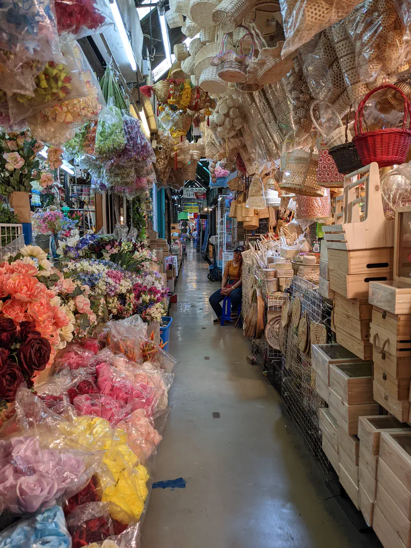 Narrow aisle in Chatuchak Market lined with artificial flowers, woven baskets, and wooden boxes for sale.