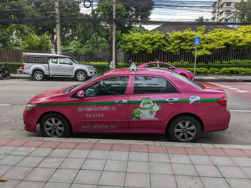 Pink and green taxi parked on a street in Thailand with a cartoon mascot on the side and Thai text.