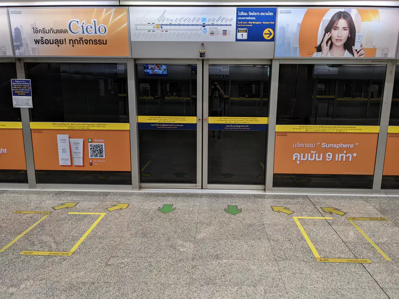 Bangkok MRT station platform with glass doors, advertisements, and marked waiting areas on the floor.