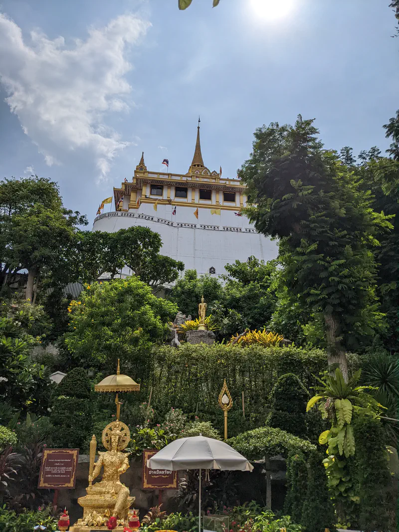 Golden Mount Temple in Bangkok viewed from below with golden statues and lush greenery under bright sunlight.