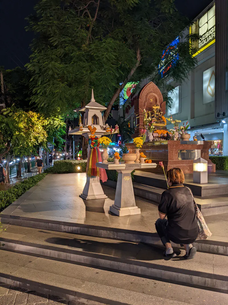 Person kneeling in prayer at a small shrine on a Bangkok sidewalk at night with offerings and flowers.