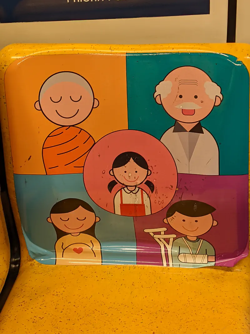 Colorful sign on a Bangkok MRT priority seat showing cartoon icons of a monk, elderly man, pregnant woman, child, and injured person.