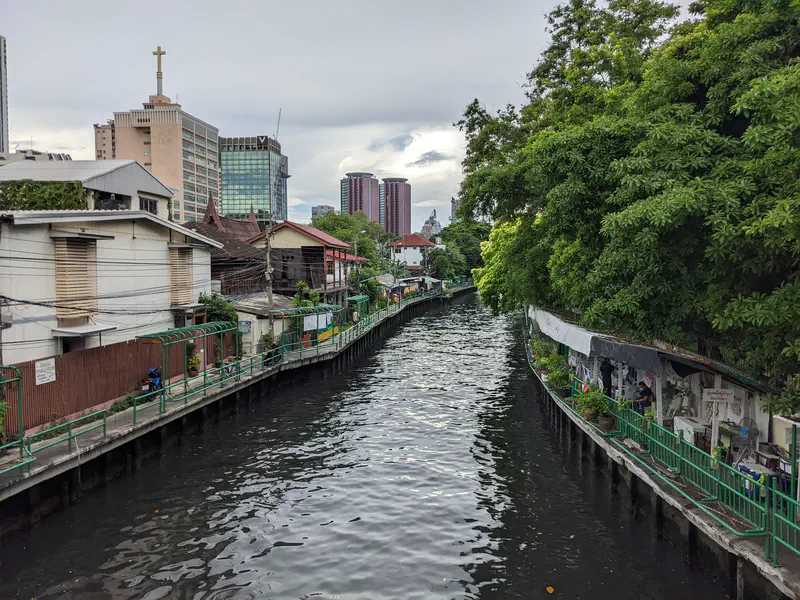 Canal in Bangkok with houses and walkways on both sides, trees overhanging the water, and modern buildings in the background.