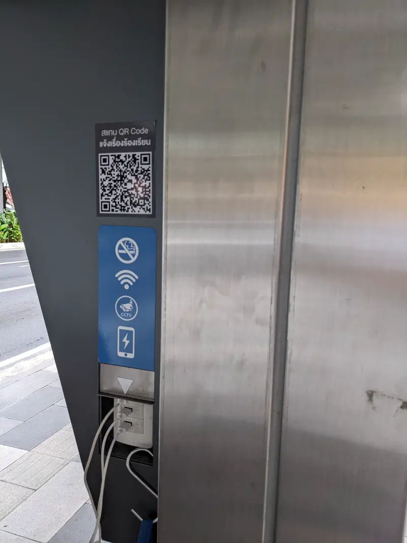 Public charging station in Bangkok with power outlets, QR code, WiFi, and CCTV signs.