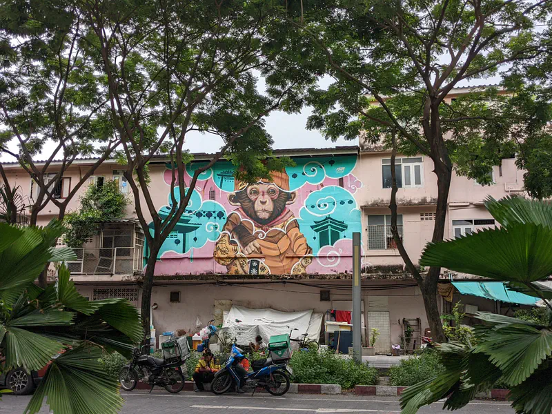 Colorful mural of a monkey in a hat painted on a building facade in Bangkok, framed by trees.