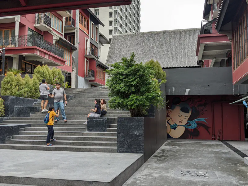 Modern courtyard with people sitting on steps and a mural of a child on the wall in Bangkok.