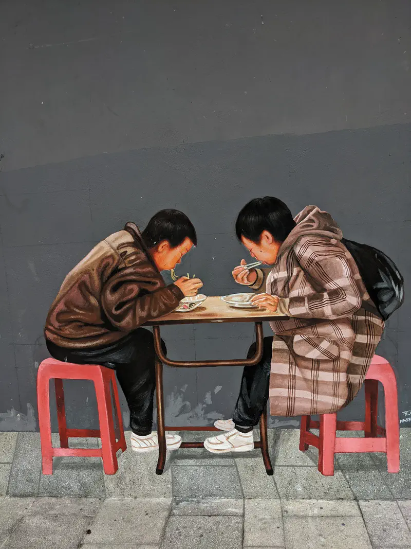Street mural depicting two people in jackets eating noodles at a small table in Bangkok.