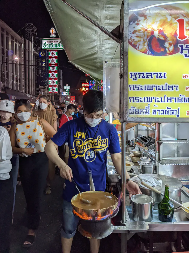 Street food vendor cooking over open flame in Bangkok Chinatown with a long line of customers.