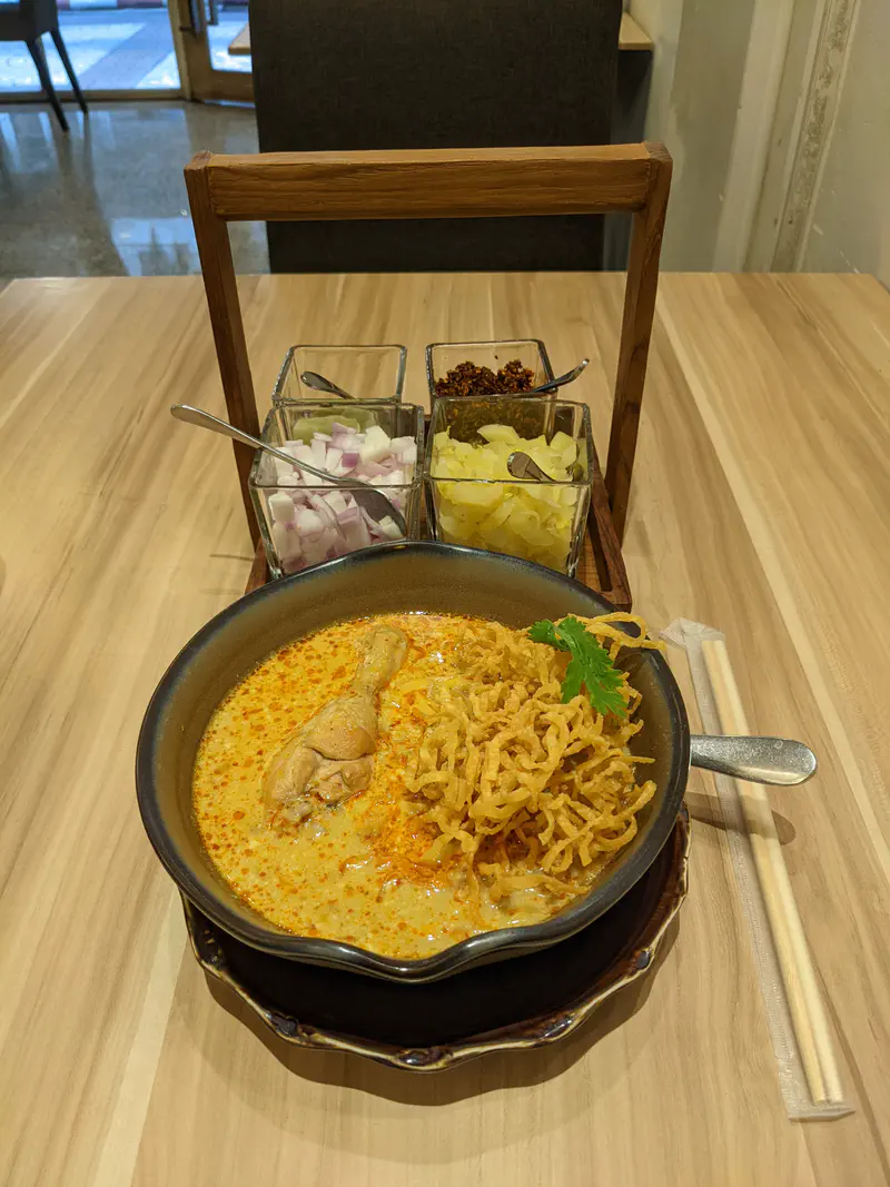 Bowl of khao soi, a Northern Thai curry noodle soup with chicken and crispy noodles, served with condiments of onion, pickled mustard greens, and chili flakes.
