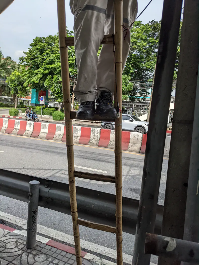 Worker standing on a bamboo ladder by a roadside, wearing safety boots and white work pants.