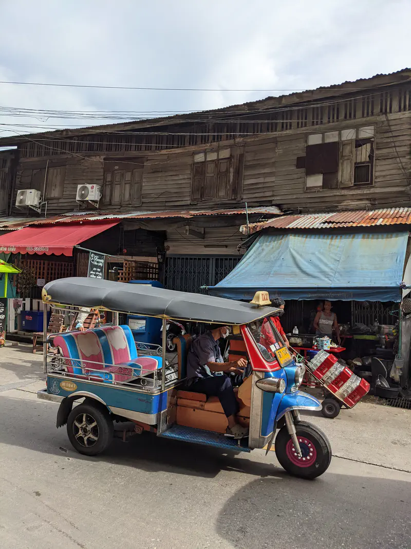 Blue and pink tuk-tuk taxi driving past old wooden buildings with corrugated roofs in Bangkok.
