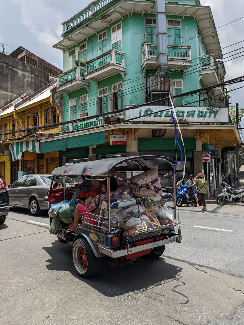 Loaded tuk-tuk taxi carrying goods and passengers passing by colorful old buildings in Bangkok.