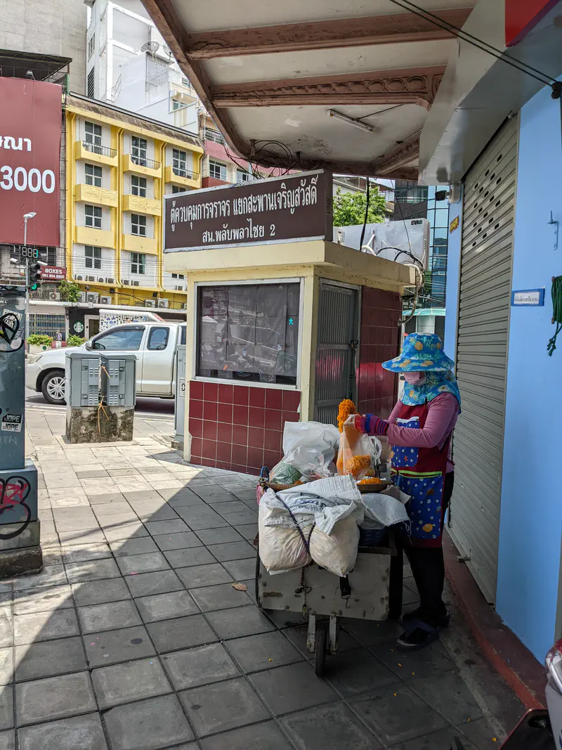 Street vendor in a blue hat and apron preparing marigold garlands at a small cart filled with bags of flowers, standing on a Bangkok sidewalk.