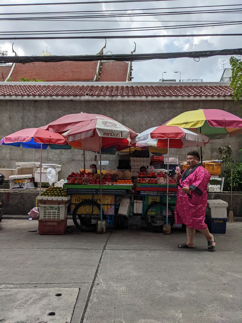 Fruit vendor stall on the street with colorful umbrellas, stacked with oranges, tomatoes, and other fruits, while a person in a pink robe walks by holding a selfie stick.